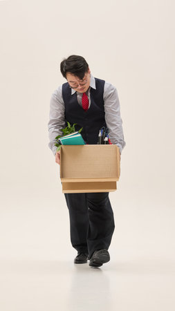 Sad Asian man dressed formal attire, holding huge cardboard box with clothes against white background. Concept of human emotions, fashion, quitting or fired job.の写真素材