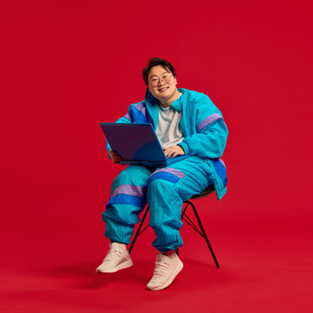 Attractive Asian man sitting on chair with laptop and looking at camera with cheerful smile against red background. Concept of human emotions, technology, digital age, working online. Adの写真素材