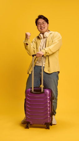 Overjoyed young Asian man, traveler clenching fist of happiness he have nice holidays, standing next to huge bag against sunny yellow background. Concept of human emotions, journey, traveler.の写真素材