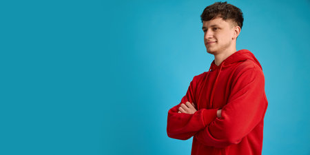 Young man in red hoodie with crossed arms looking to side with smirk against blue studio background....