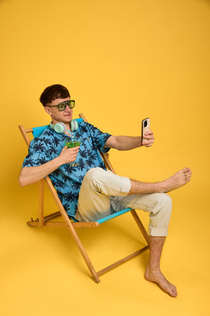 Full body photo of smiling man taking selfie in deck chair with green drink, dressed for beach holiday. Concept of social media, relaxation, lifestyle, vacation, content creation, leisure.の写真素材