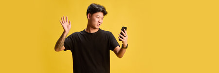 Banner. Cheerful young man waving at smartphone while making video call, standing on yellow...