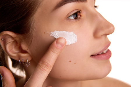 Close up portrait of young woman applying rejuvenating, nutrient cosmetic product on cheeks against...