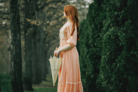 Young woman with auburn hair in delicate vintage dress stands still near tall firs, holding ivory umbrella, lost in silence. Concept of solitude, fantasy, nostalgia. Suitable for book or movie cover.の写真素材