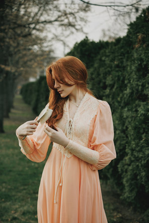 She brushes her copper strands with ritual calm, lost in gesture, her peach gown glowing against deep green. Concept of self-reflection, femininity, ritual. Suitable for book or movie cover.の写真素材