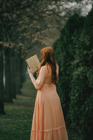 Back turned, book held aloft, she reads among trees like ghost of knowledge haunting forgotten pages. Concept of solitude, memory, intellectual passion. Suitable for book or movie cover.の写真素材