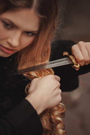 Close-up of fearless young woman cutting long red curls with ornate dagger. Fantasy characters. Concept of transformation, revenge, symbolic sacrifice. Suitable for dark fairytale or drama.の写真素材