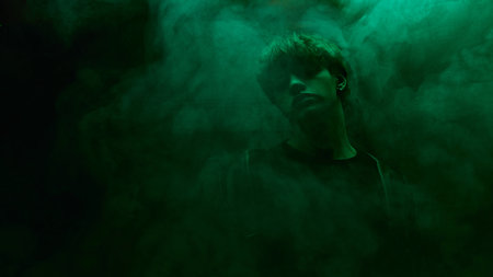 Young man in dark shirt and leather coat stands surrounded by green mist in moody atmosphere against dark studio background. Concept of beauty and fashion, self-expression, human emotions.の写真素材