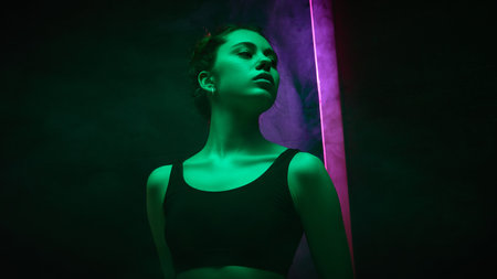 Young woman in green-pink glow stares seriously with subtle turn against gradient pink-dark background with vertical light. Expressive portrait. Concept of beauty and fashion, self-expression, emotionの写真素材