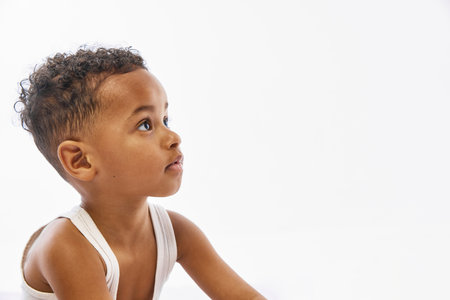 Close up photo of curious toddler boy in white tank top looks up with calm expression against white studio background with negative space. Concept of beauty, childhood, parenthood, wellness.の写真素材
