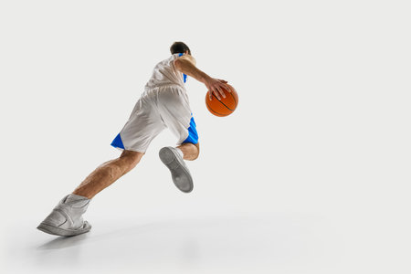 Low angle view. Young basketball player runs forward with determination, gripping ball tightly and dressed in game uniform against white background. Concept of focus, sport events, energy, competitiveの写真素材