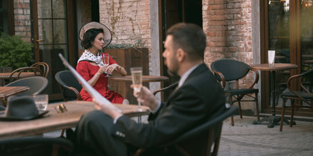 Vintage couple in love. Woman in red dress looks at man reading paper while holding champagne,...