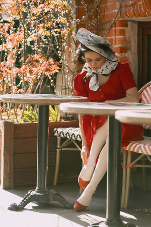 Graceful and elegant woman in red dress adjusts her shoe while sitting in morning light between cafe...