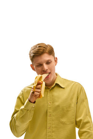 Young man in green shirt biting ripe banana with playful face and closed eyes showing joy and appetite in expressive motion against white studio background.の写真素材