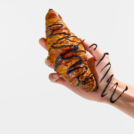 Hand gripping chocolate-drizzled croissant with syrup messily swirling down wrist against white studio background. Concept of fast food, takeaway, catering, traditional, nutrition, lunch.の写真素材