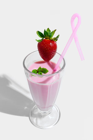 Tall glass of pink strawberry smoothie topped with fresh whole strawberry, green mint leaf and curly straw serving against white studio background. Concept of nutrition, takeaway, catering.の写真素材