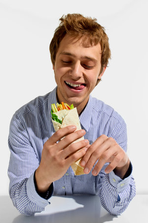 Happy young man, in striped shirt, holding vegetable wrap with playful tongue out, enjoying colorful healthy snack against white background.の写真素材