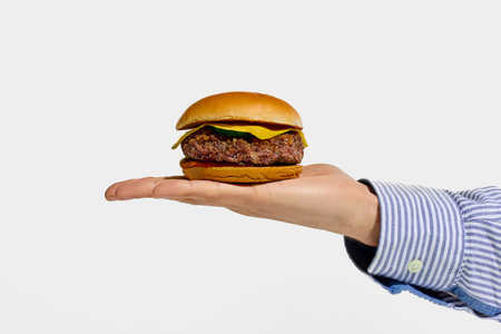 Hand presenting juicy cheeseburger with beef patty, pickles and melted cheese on palm against white background. Concept of fast food, takeaway, traditional, catering, nutrition.の写真素材
