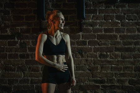 Athletic woman standing relaxed in gym smiling confidently after exercise with hand resting on abs, showing fitness and self-pride against brick wall. Concept of sport, active and healthy lifestyleの写真素材