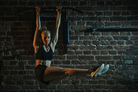 Woman lifting straight legs while hanging from bar in gym performing challenging core exercise with control, strength and balance. Concept of sport, active and healthy lifestyle, competition.の写真素材