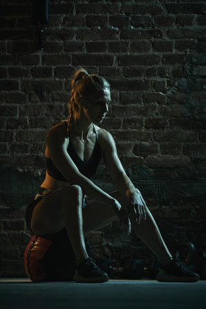 Fit woman sits on medicine ball catching breath after intense workout session, muscles glistening as she focuses under low dramatic lighting. Concept of sport, active and healthy lifestyle, trainingの写真素材