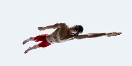 Focused male swimmer in red shorts, goggles and cap extending arm in freestyle pose, muscles tensed in suspended motion against white backdrop. Concept of sport, training, discipline, motionの写真素材
