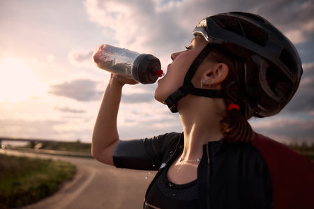Close-up photo of female cyclist pouring water from bottle into mouth, sunlight behind and road curving in distance, drops splashing in midair. Concept of refreshment, heat, moment, strength, balance.の写真素材