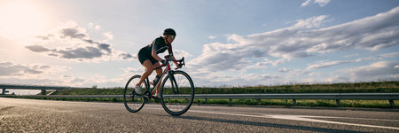 Wide angle view photo of cyclist on open highway, riding under dramatic sky with bridge in distance and warm evening light casting soft. Concept of extreme sport, fitness, road cycling, freedom, speedの写真素材