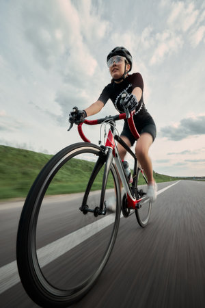 Wide angle. Woman on racing bike with clear expression of focus, riding along clean asphalt road,...