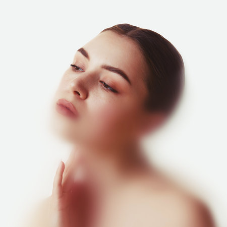 Conceptual portrait. Woman tilts head gently with hand near neck in diffuse white light, emotion of gentle introspection and vulnerability. Concept of femininity, emotion, softness, self-care.の写真素材