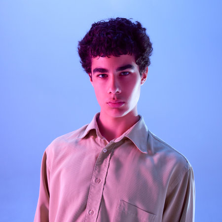 Teen boy with curly hair in beige shirt confidently looking straight, illuminated by pink and blue...