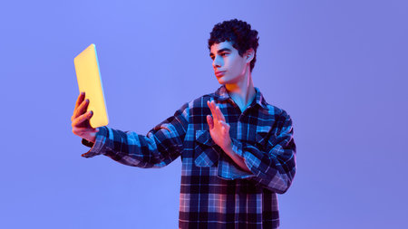 Stylish teenager posing with tablet in hand, looking to side as if recording video message in neon...
