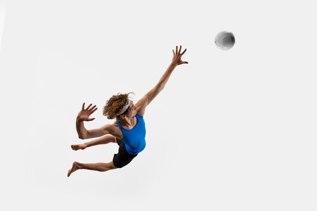High angle view of jumping athlete extends one hand high above in dynamic motion as volleyball approaches against white studio background. Concept of sport, championship, powerの写真素材