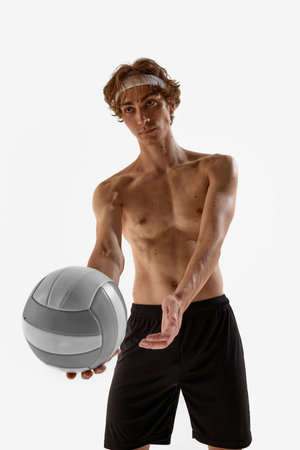 Young male athlete in black shorts and headband stretches hand forward offering volleyball with serious expression against white background. Concept of sport championship, victory, wellness, energyの写真素材