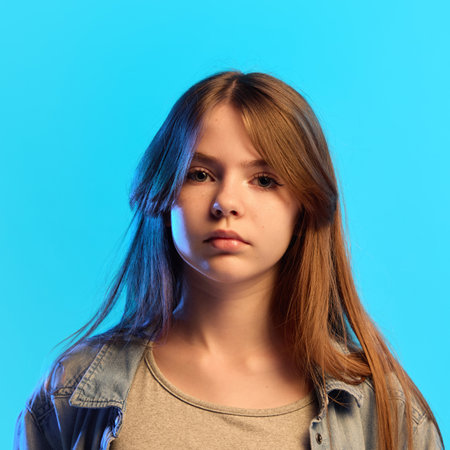 Close-up portrait of smiling young teen girl dressed casual attire looking at camera cheerfully in neon light against blue studio background. Concept of human emotions, youth, fashion, calmnessの写真素材