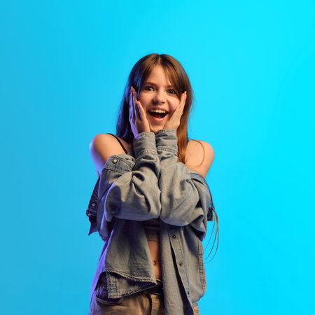 Pretty teen girl holds hands to cheeks in amazed emotion with wide eyes under blue neon light in...