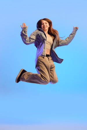 Teen girl jumping in air with joyful face and arms raised in casual clothes against blue background...