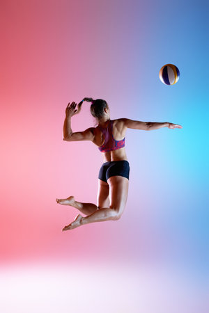 Photo in motion, rear view of jumping athlete with extended arm targeting volleyball in neon split-color light against gradient background. Concept of strength, accuracy, and momentum in sport.の写真素材