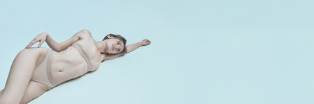 Banner. Model in beige underwear lies on side with arm extended, soft light casts gentle shadows on skin against pale blue background with copy space. Concept of modern beauty, calmness, self-careの写真素材