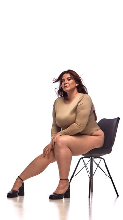 A plus-size woman confidently poses on a chair in an editorial style outfit with bold visual contrast. Concept of body positivity and fashion diversity.の写真素材