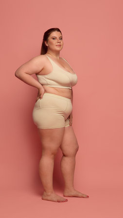 A confident woman embraces natural body diversity, posing in comfortable nude-toned underwear against a pink backdrop. Concept of body positivity and self-acceptance.の写真素材