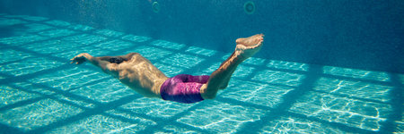 A person swims underwater in a pool as sunlight creates a grid pattern on the clear blue water....