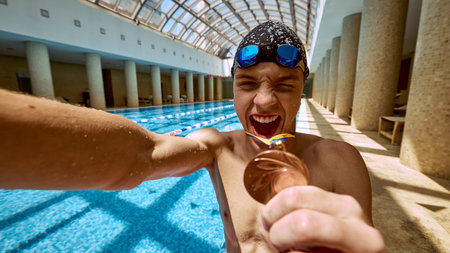 A young swimmer celebrates victory holding a medal in an indoor pool, featuring an arched glass ceiling and sandy columns. Concept of achievement and inspiration in sports.の写真素材