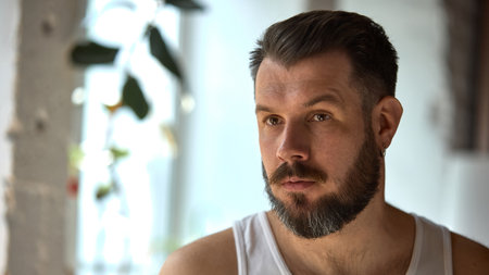 Skin-realism. Relaxed man with beard wearing white tank top sits indoors. Defined cheekbones highlighted with side lighting, no makeup. Concept of peaceful solitude and comfort at home.の写真素材