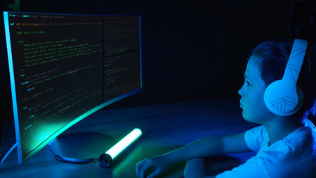 Side view of a boy with headphones looking at code on a large curved screen in a neon blue-lit room....