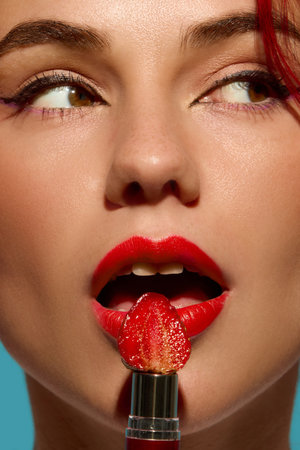 Closeup of woman lips painted red with strawberry on lipstick tube against teal background. Concept...
