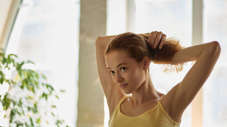 Clean-lazy hybrid. Woman with focused expression tying back hair while getting ready in morning...