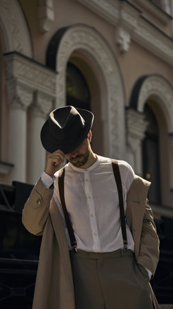 Man in vintage attire tips his hat while standing in front of an ornate building with arched...
