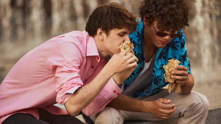 Two young men eating shawarma on city steps in casual vibrant clothing. Concept of carefree summer moments, food delivery promotion, fast food marketing, modern social habits.の写真素材