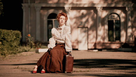 Woman in white blouse and long skirt sitting on pavement with suitcase and red shoes. Concept of...